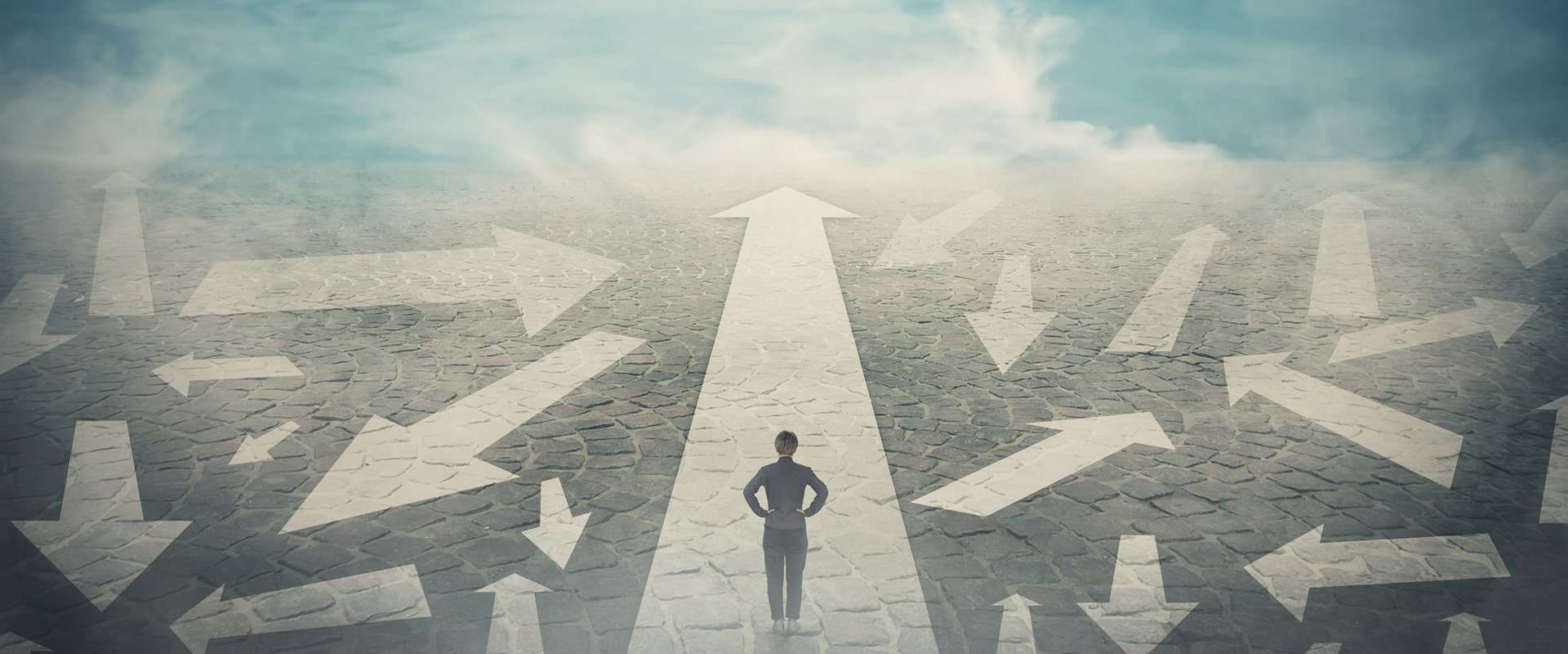 Doubtful person, hands on hips, choosing the way as multiple arrows on the road showing a mess of different directions. Choosing the correct pathway, difficult decision concept, confusion symbol. (StunningArt/Shutterstock.com)